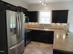 A kitchen with black cabinetry features a stainless steel refrigerator, an electric stove, a microwave, and a double sink. The countertops are brown, complementing the tile flooring. A small plant sits on the counter, and a window provides natural light.