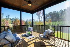 A screened-in porch with a wooden floor features a ceiling fan and three cushioned chairs around a low table. The space overlooks a sunny, scenic view of trees and a grassy area in the background.
