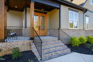 Front entrance of a house with a wooden and glass door, brick steps, and a small porch. The exterior features beige siding and brickwork. Black railings line the steps and porch, with a small seating area to the left and shrubs along the front.