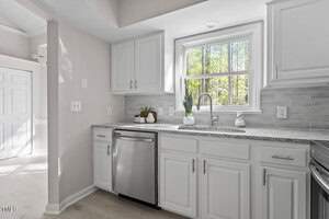 A modern kitchen with white cabinets and a gray tile backsplash. It features a stainless steel dishwasher and a window above the sink, letting in natural light. The countertop has a potted plant and other small items. Light-colored flooring is visible.