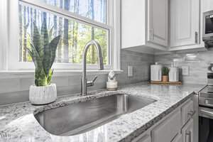 A modern kitchen sink with a granite countertop and stainless steel faucet beneath a bright window. There is a potted plant and a dispenser on the counter. The backsplash is gray tile, and two white storage jars are on a wooden tray.
