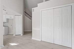 A modern, empty room with light-colored hardwood floors and white walls. The room features a tall ceiling, a stairway, and double doors. A small section of a kitchen with closed cabinets is visible on the left. The space is well-lit with natural light.