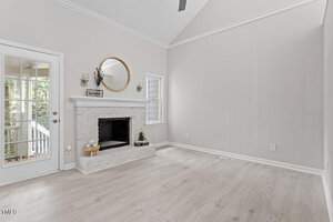 A minimalist living room with light-colored walls and wooden flooring features a white brick fireplace. Above the mantel hangs a round mirror. A door with glass panes and a small window can be seen on the left side, letting in natural light.