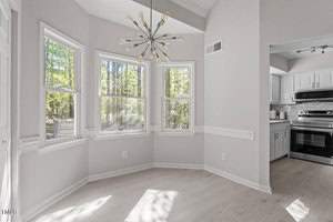 A bright dining area with large windows overlooking a wooded scene. The room features light-colored wood flooring and a modern chandelier. In the background, there's a view of a kitchen with stainless steel appliances and white cabinetry.
