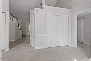 An empty room with light-colored hardwood floors and white walls. The space includes a white double-door closet and a staircase with a wooden railing. A windowed door leading outside is visible in the background. Natural light illuminates the area.