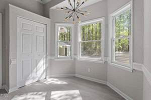 A bright room with gray walls and light-colored laminate flooring. Large windows provide a view of trees outside. A modern chandelier hangs from the ceiling. A closed white door is on the left. Sunlight casts shadows on the floor.