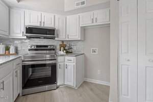 Modern kitchen with white cabinets, silver appliances including a stove and microwave, and a marble-like backsplash. A wooden cutting board and plant are on the counter. There are white folding doors on the right and light wood flooring throughout.