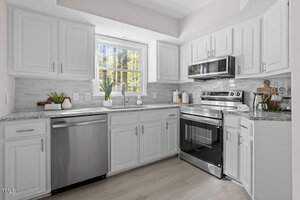A modern kitchen with white cabinets, stainless steel appliances, and a gray tile backsplash. A window above the sink provides natural light. The countertop features decorative plants and kitchen utensils. Light wood flooring complements the space.