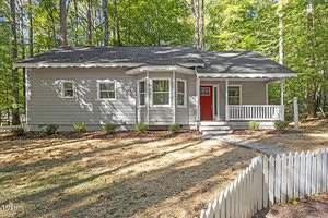 Located at 3080 Willow Creek Drive in Wake Forest, this charming single-story gray house boasts a red front door and a cozy porch, nestled among trees. Several windows brighten the view, while a white picket fence and landscaped shrubs add to its inviting appeal.