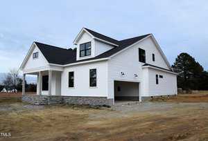 A modern two-story white house at 1590 Nc 96 Hwy in Franklinton boasts black roof accents and stone detailing around the base. Situated on a cleared plot with dirt and sparse grass, this home features multiple windows and an attached two-car garage.