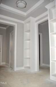A room with gray walls and white trim features a built-in corner bookshelf. The ceiling has tray molding and a modern circular light fixture. The floor appears unfinished, with visible areas of cement. Multiple doorways are present.