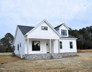 A newly constructed, white modern farmhouse-style house with a gabled roof. It features black-framed windows, a stone foundation, and a simple front porch. The house is set on a mostly bare plot with sparse grass and trees in the background.