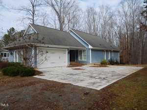 The single-story house at 513 Port Drive, Henderson, features pale blue siding and a gray roof, surrounded by bare trees. It boasts a double garage and a wide driveway. The mostly brown yard hosts some shrubs near the house, set under an overcast sky.