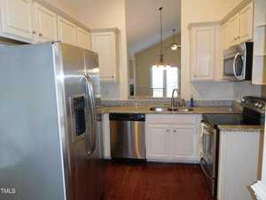 A kitchen with stainless steel appliances including a refrigerator, dishwasher, and oven. Light-colored cabinets and speckled granite countertops are present. A double sink is under a window with vertical blinds. The floor is hardwood.