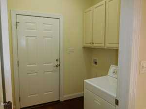 A laundry room with a white washing machine on the right, beige cabinets above, and a closed white door on the left. The walls are painted light yellow, and the floor is dark wood.