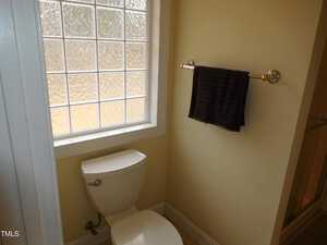 A small bathroom corner features a white toilet beneath a frosted glass window. A dark towel hangs on a silver towel rack mounted on a yellow wall. The floor is tiled.