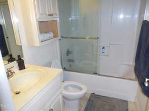 A small bathroom with a white toilet, sink, and cabinet. A glass sliding door encloses the bathtub and shower. A beige mat is on the floor, and a navy towel hangs to the right. A roll of toilet paper and a bottle are on the cabinet shelf.