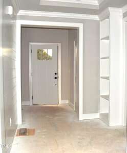 A hallway with light gray walls and a white front door. The floor is unfinished, showing plywood, and there are built-in white shelves on the right. Natural light comes through the door's glass panel.