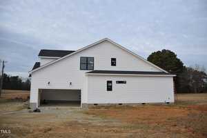 A newly constructed white house with a dark roof and green windows stands on a cleared lot. The garage is open, and a ladder leans against the structure. The surrounding area is mostly bare ground with a few trees in the background.