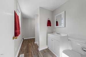 A bathroom with laminate wood flooring, a white vanity with a mirror, and a white toilet. Two red towels hang on the walls. The room is painted light gray, and there is natural light coming in from a window.