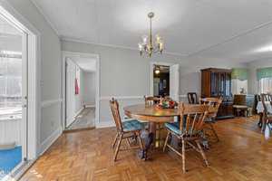 A dining room with a wooden table and five chairs on a parquet floor. A chandelier hangs above the table, which has a floral centerpiece. A china cabinet is in the corner. A hallway door is open, revealing an adjacent room. Natural light fills the space.
