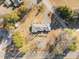 Aerial view of a suburban area featuring a large house with a gray roof surrounded by trees and a lawn. A winding driveway connects the house to a road. Other houses and roads are visible in the surrounding landscape.