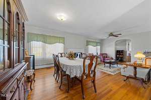A dining room with a wooden table covered by a lace tablecloth, surrounded by chairs. A cabinet is on the left, while a living area with armchairs and a rug is visible in the background. The room has hardwood floors and large windows with green valances.