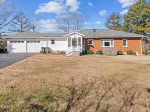 Single-story brick and white panel house with a two-car garage, front porch, and a large, grassy yard. A paved driveway leads to the garage. The sky is partly cloudy, and trees border the property.