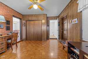 A room with wood-paneled walls and parquet flooring features a ceiling fan with a light. There's a wooden desk with a chair on the left and a doorway leading to the dining area. Various decor items and plaques are mounted on the walls.