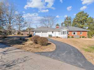 A single-story house with a gray roof and red brick exterior is seen on a sunny day. A curved driveway leads to a two-car garage. The yard is mostly grass, bordered by trees and shrubs. The sky is partly cloudy.