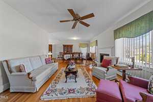 A living room with a floral rug, sofa, and two cushioned chairs. There's a wooden coffee table with flowers, a ceiling fan, and a dining table set in the background. Large windows provide natural light and green drapes adorn the windows.