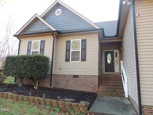 A beige house with a dark gray roof and a brick foundation. There are black shutters on the windows and a wooden door with an oval glass insert. Bushes line the left side of the entrance, and a small concrete path leads to the steps.