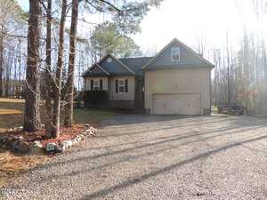 A tan, two-story house with a garage is situated in a wooded area. It features a gable roof, a small front porch, and a gravel driveway. Leafless trees surround the property, and the sunlight creates a bright atmosphere.