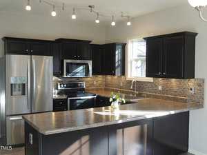 Modern kitchen with dark cabinets, a stainless steel refrigerator, and a built-in oven with a microwave above. The countertop is L-shaped with a single plant decoration. The backsplash features small, rectangular tiles, and ceiling lights provide illumination.