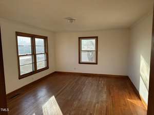 An empty room with hardwood floors and white walls. Two large windows on the left wall and one window on the back wall allow natural light to enter. A ceiling light fixture is centered on the ceiling.
