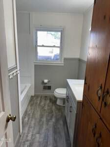 A small bathroom featuring a white bathtub with a shower, gray flooring, a white toilet, and a vanity with a sink. A wooden cabinet is on the right, and a window is above the toilet, allowing natural light. The walls are white with gray wainscoting.