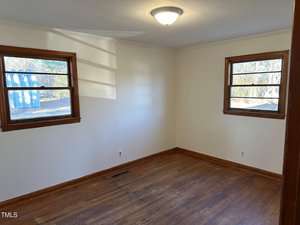 An empty room with white walls and hardwood flooring features two wooden-framed windows on adjacent walls. A ceiling light fixture is centered on the white ceiling. Sunlight streams in through the windows, casting shadows on the wall.