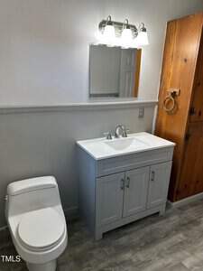 A bathroom with a modern white toilet and a gray vanity with a sink. A simple mirror and a light fixture with four bulbs are mounted on the wall above the sink. A wooden cabinet is on the right side. The floor has a wood-like finish.
