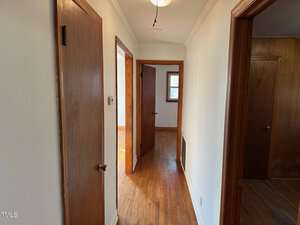 A hallway with hardwood floors and white walls, featuring three wooden doors on the left and right. A ceiling light fixture is visible. Sunlight filters through a small window at the end of the hallway.