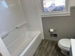 A bathroom with a white bathtub and a toilet on a wood-patterned floor. There is a wall-mounted toilet paper holder and a window showing a parked truck outside. The walls are light gray with white trim.