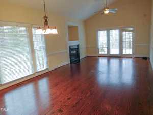 A spacious living room with wooden flooring and cream-colored walls. Large windows with blinds allow natural light. A hanging light fixture is above, and a ceiling fan is on the high ceiling. French doors open onto a terrace.