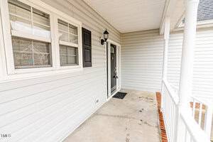 A residential front porch with light gray siding and a black front door. There is a light fixture next to the door. The porch has a concrete floor and a white railing. A welcome mat is placed at the entrance.
