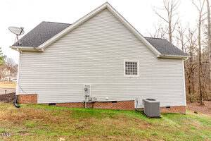 The image shows the back of a single-story house with white siding and a black roof. It has a small window, a satellite dish, and an air conditioning unit. The house is bordered by grass and situated near a wooded area.