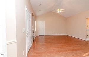 A spacious, empty room with light brown hardwood floors and beige walls. The ceiling is vaulted with a white ceiling fan. There are two white doors on the left, and a small cutout window on the right wall.