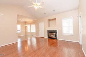 Spacious living room with hardwood floors, a ceiling fan, and a fireplace. There are two large windows with blinds and an open doorway leading to a dining area with a chandelier. Walls are painted in a light neutral color.