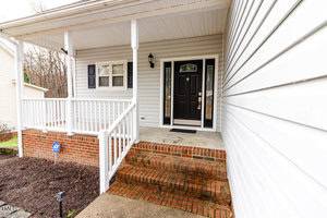 The image shows the front entrance of a house with a brick staircase leading up to a covered porch. The porch features white railings and a black front door with sidelights. The house siding is white, and there is a small yard with a security sign.