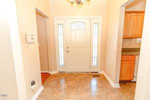 A well-lit entryway with a white front door featuring glass panels and sidelights. The floor is tiled, and the walls are painted in a soft beige. A thermostat is mounted on one wall, and wooden kitchen cabinets are partially visible on the right.