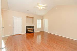 A spacious living room with light-colored walls and hardwood flooring. The room features a ceiling fan, a fireplace with a stone surround, and two doors. There is a window on the right, allowing natural light to enter.