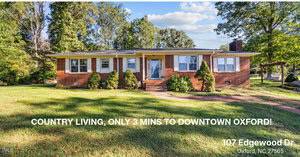 A charming one-story brick house at 107 Edgewood Drive, Oxford, with a well-maintained lawn and shrubs. Nestled among tall trees and beneath a partly cloudy sky. Experience country living just 3 minutes from downtown!.