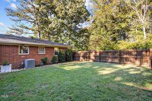A brick house with a backyard featuring a lawn and a wooden privacy fence. A small air conditioning unit is visible next to the house. Tall trees border the fence, and the sky is clear with a few clouds.